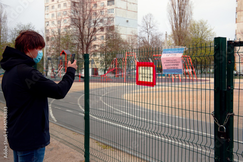 Kharkov, Ukraine, 18.04.2020:
Guy in mask look at warning 
sign of quarantine by covid-19 on 
iron fence of closed kids playground with tied of white and red boundary tape in carantin time  photo