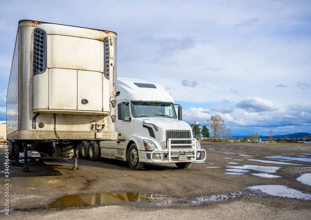 Big rig semi truck with grille guard standing side by side with refrigerated semi trailer on the with parking lot with puddles waiting for the next load for delivery
