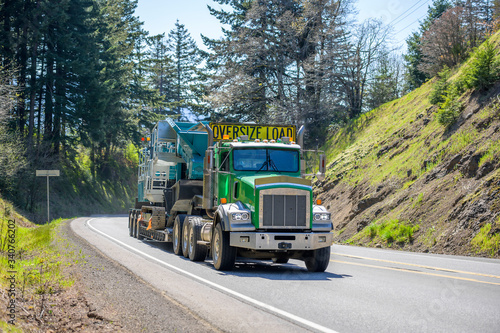 Green powerful big rig semi truck with oversize load yellow sign on the roof transporting crawler on the step down semi trailer running uphill on the winding mountain road