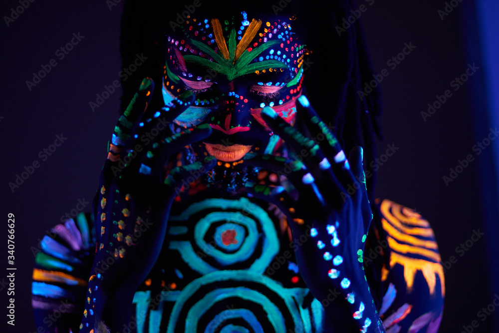 Naklejka premium fantastic mysterious african male with UV body art posing at camera, futuristic body art, fluorescent paints glows in neon light