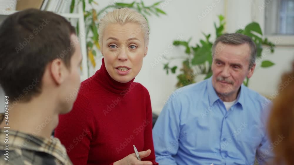 Tilt up medium shot of smiling middle aged female psychologist with clipboard talking to patients during group therapy session Tilt up medium shot of smiling middle aged female psychologist with clipboard talking to patients during group therapy session