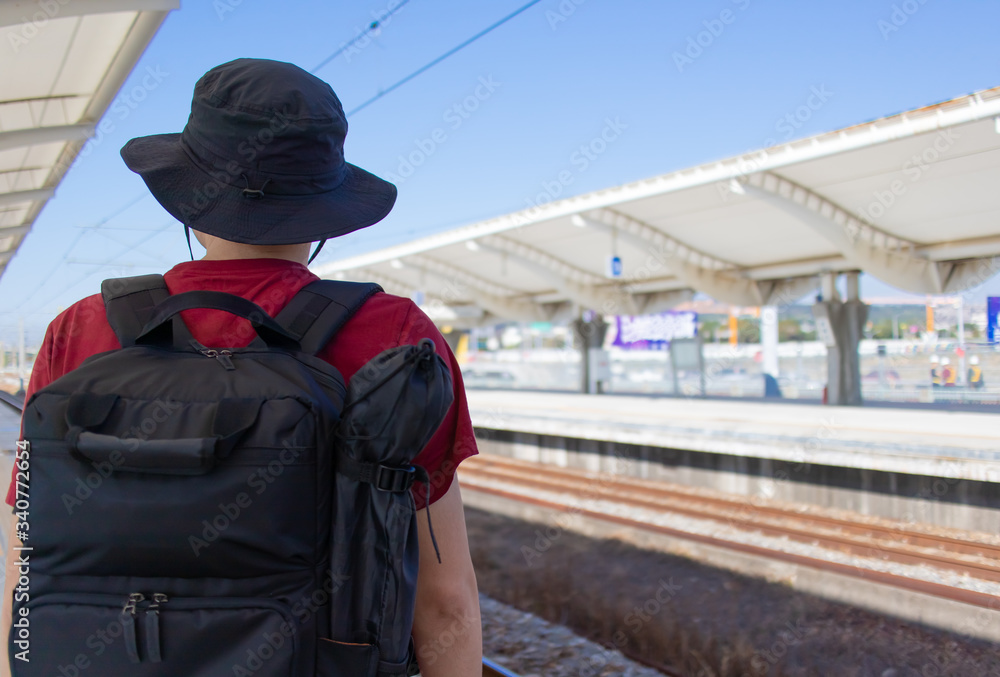 鉄道の駅のプラットフォームで電車を待つ黒いリュックサックを背負った男性の後ろ姿 鉄道の駅のプラットフォームで電車を待つ黒いリュックサックを背負った男性の後ろ姿