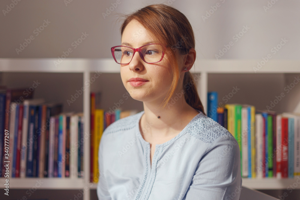 Front view portrait of beautiful young adult caucasian woman female girl sitting in the chair at home or office in front of the bookshelf looking to the side wearing eyeglasses shirt in day Front view portrait of beautiful young adult caucasian woman female girl sitting in the chair at home or office in front of the bookshelf looking to the side wearing eyeglasses shirt in day