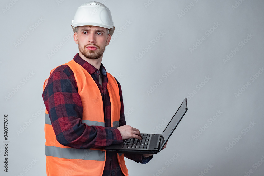 A man in an orange vest and hard hat is holding a laptop. Construction engineer. The architect in the hard hat looks away. Monitoring the progress of construction. Checking all stages of construction. A man in an orange vest and hard hat is holding a laptop. Construction engineer. The architect in the hard hat looks away. Monitoring the progress of construction. Checking all stages of construction.