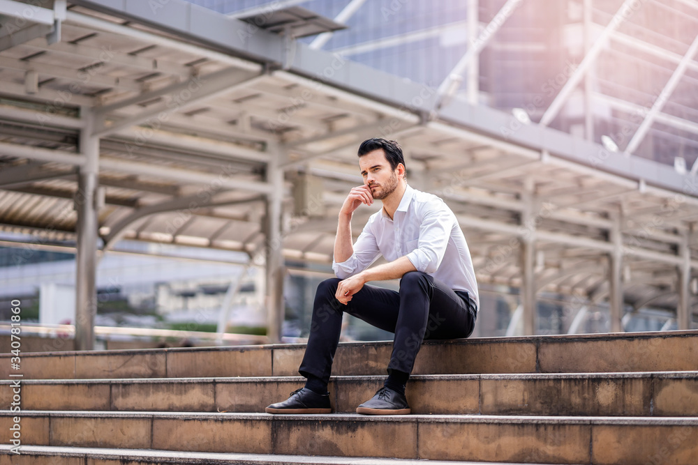 male business worker sitting on top of stairway with hand against his cheek thinking, representing getting brain storming or stressed out from hard work, within a modern city urban structure district