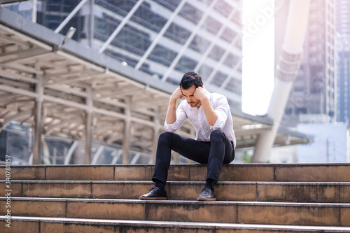 male business worker sitting on top of a stairway with fists against head looking down feeling sad, representing getting fired from a job or stressed out from hard work, within an urban city district