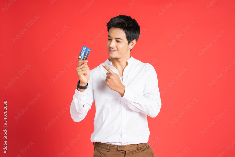 attractive asian businessman smiling holding pointing finger at debit card of financial payment concept, wearing casual clothing and black earrings, standing and posing with red isolated background