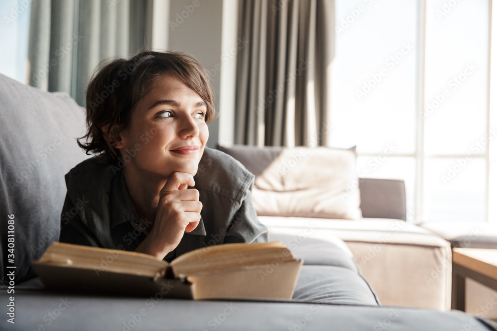 Image of caucasian cute pleased woman reading book and smiling - obrazy, fototapety, plakaty 