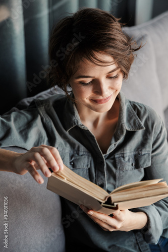 Image of brunette cute pleased woman reading book and smiling