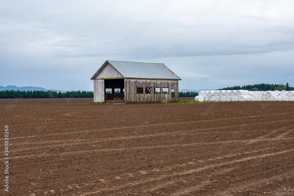 An old, weathered wood shed with a metal roof and open ends sits at the edge of a tilled field of dirt in Oregon, with white plastic covered rolls of hay lined up in the background. 