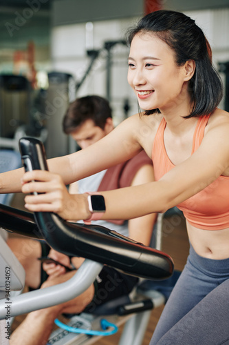 Happy young woman riding on spinning bike when her boyfriend or fitness coach sitting nearby and checking text messages