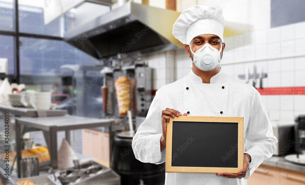 health protection, safety and pandemic concept - indian male chef cook wearing face protective mask or respirator with blank chalkboard over kebab shop kitchen background