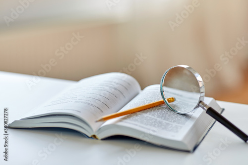 education, school and learning concept - book with magnifier and pencil on table at home