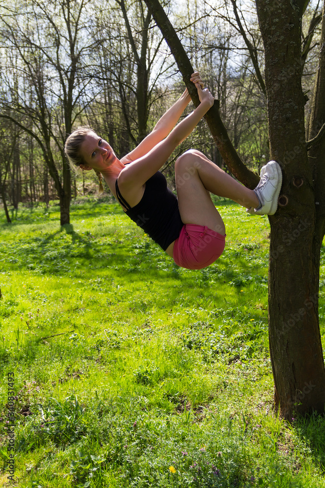 Portrait of young blonde caucasian woman climb on spring tree in avenue with grass Portrait of young blonde caucasian woman climb on spring tree in avenue with grass