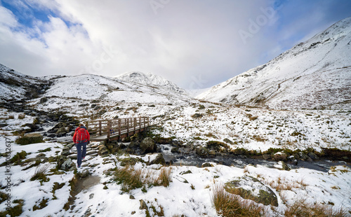 A hiker crossing Glenridding Beck over a wooden bridge on route to a snow covered Catstye Cam and Helvellyn in the English Lake District.