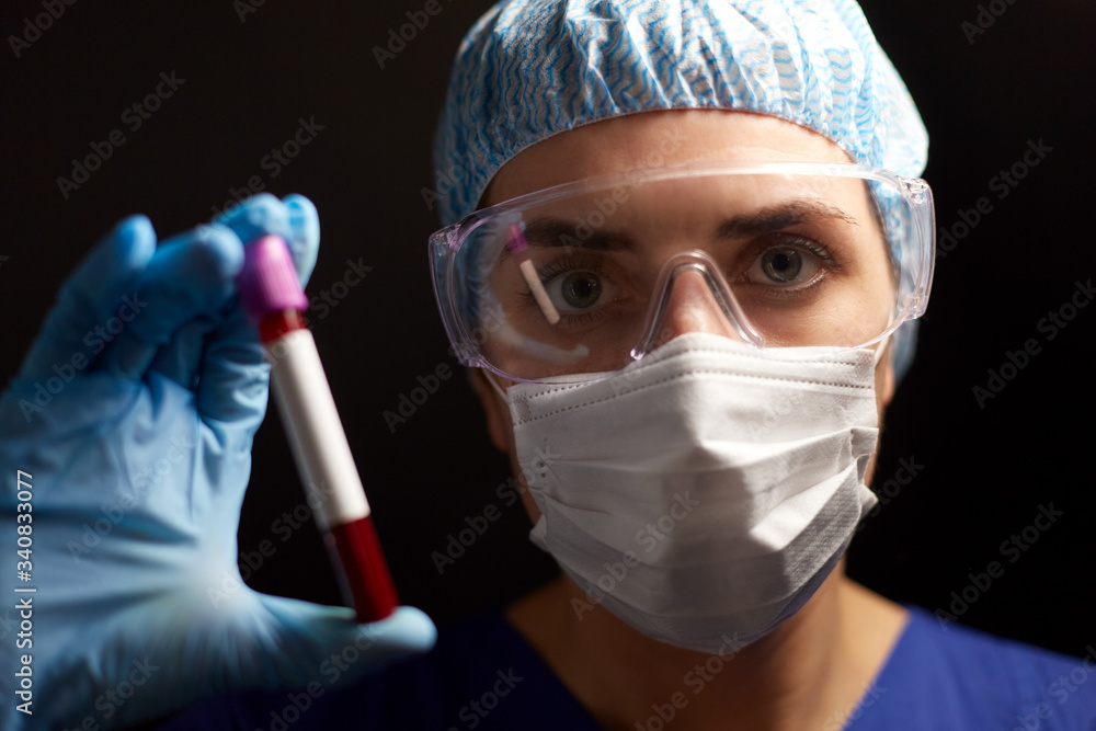 medicine, health and pandemic concept - close up of young female doctor or nurse wearing goggles, glove and face protective mask holding beaker with blood test over black background