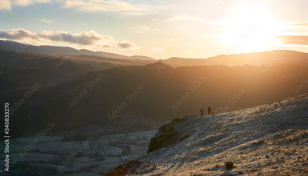 Two hikers watching the bright orange sunny winters morning sunrise over Borrowdale from below Maiden Moor in the Lake District, UK.