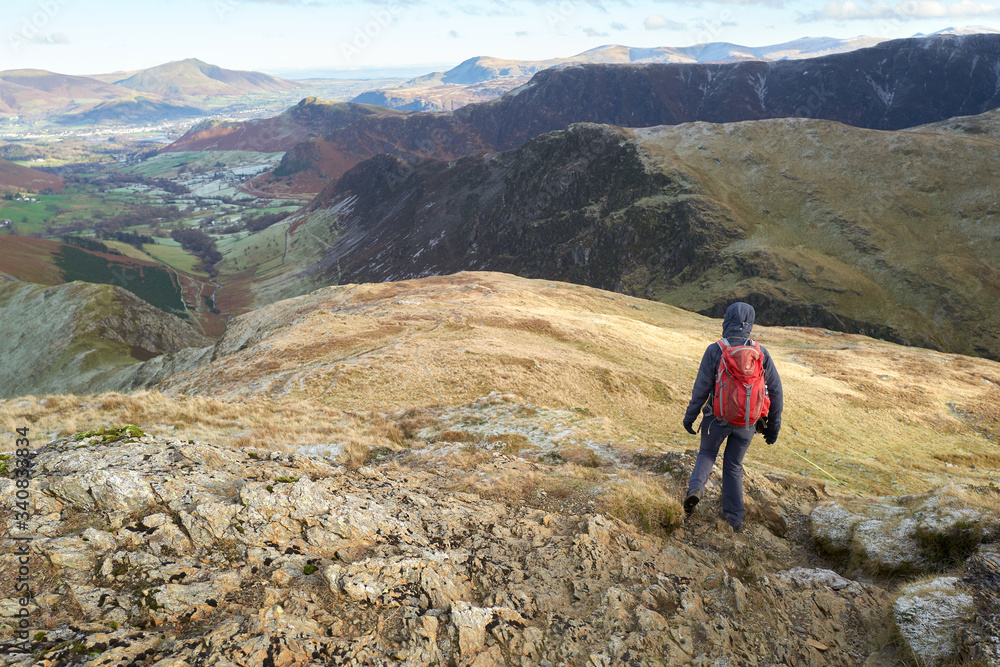 A hiker descending the rocky mountain ridge from the summit of Robinson towards High Snab Bank on a cold winters day in the Lake District, UK.