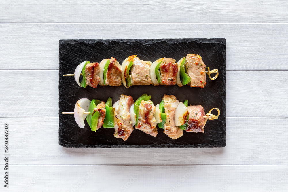Two portions of grilled pork with onion and green pepper. Barbeque with vegetables, skewers with kebab on black stone plate, on white wooden table, top view. Two portions of grilled pork with onion and green pepper. Barbeque with vegetables, skewers with kebab on black stone plate, on white wooden table, top view.