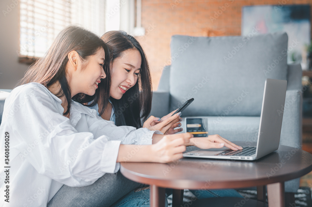 Asian women friends searching for online shopping in smartphone and laptop while holding credit card in living room Asian women friends searching for online shopping in smartphone and laptop while holding credit card in living room