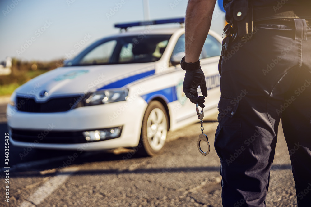 Police officer holding handcuffs, police car in background. Police officer holding handcuffs, police car in background.