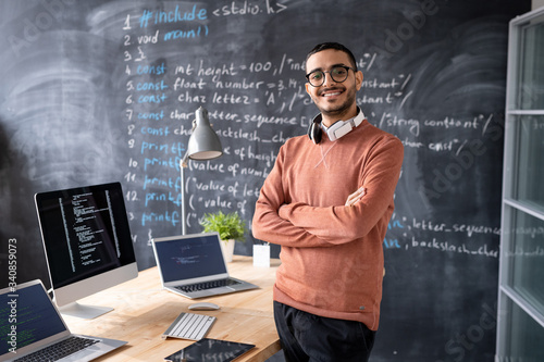 Portrait of satisfied young middle-eastern computer programmer with headphones around neck standing with crossed arms in coding studio