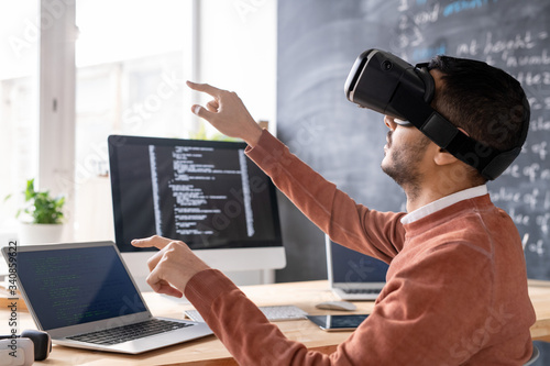 Concentrated young Arabian man sitting in computer office and developing app in VR goggles