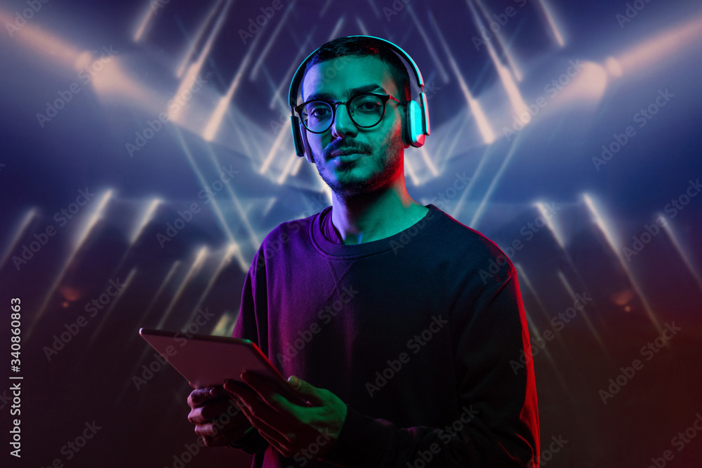 Portrait of serious middle-eastern man in wireless headphones using digital tablet against luminous background with geometric shapes Portrait of serious middle-eastern man in wireless headphones using digital tablet against luminous background with geometric shapes