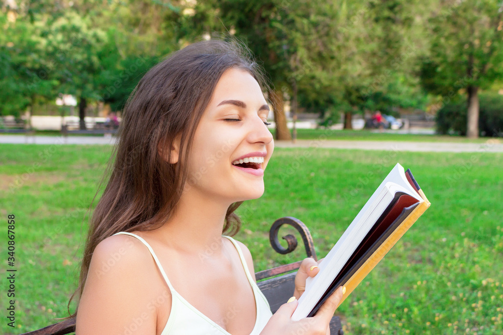 Beautiful young brownhair girl smells a fresh printed book sitting on the bench in the park Beautiful young brownhair girl smells a fresh printed book sitting on the bench in the park
