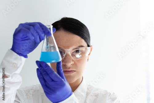 female scientist with protective eyeglasses and gloves holding a flask with blue liquid substance