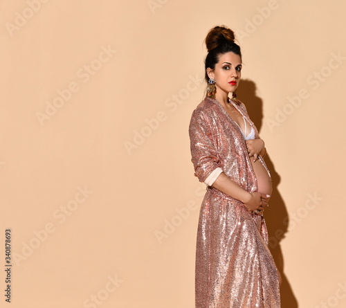 Pregnant girl in earrings, white lingerie and golden robe. She hugging her tummy, posing sideways on beige background. Close up