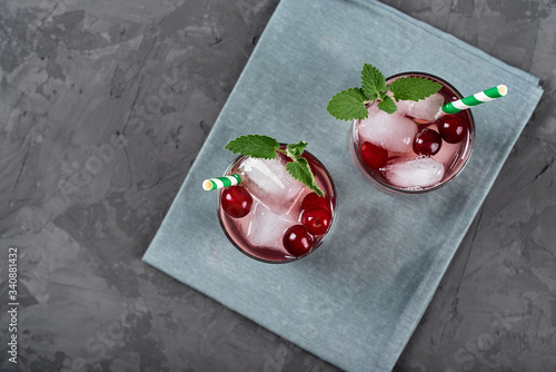 Fresh cherry lemonade with ice, mint and paper straw in sparkling glasses on gray table background, copy space. Cold summer drink. Berry cocktail