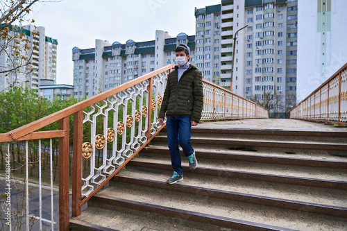 teen boy walks down the street during the day, a pedestrian walkway and high-rise buildings with apartments, a residential area, a medical mask on his face protects against viruses and dust