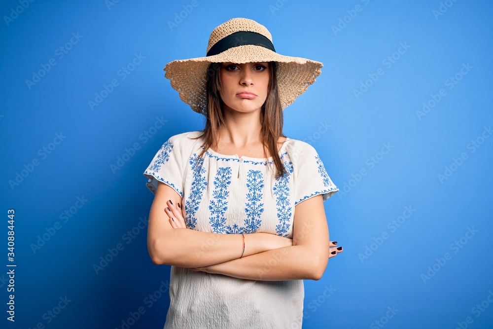Young beautiful brunette woman wearing casual t-shirt and summer hat over blue background skeptic and nervous, disapproving expression on face with crossed arms. Negative person.