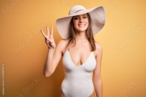 Young beautiful brunette woman on vacation wearing swimsuit and summer hat showing and pointing up with fingers number three while smiling confident and happy.