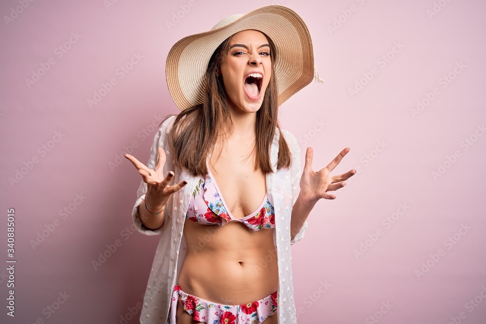 Young beautiful woman on vacation wearing bikini and summer hat over pink background crazy and mad shouting and yelling with aggressive expression and arms raised. Frustration concept. Young beautiful woman on vacation wearing bikini and summer hat over pink background crazy and mad shouting and yelling with aggressive expression and arms raised. Frustration concept.