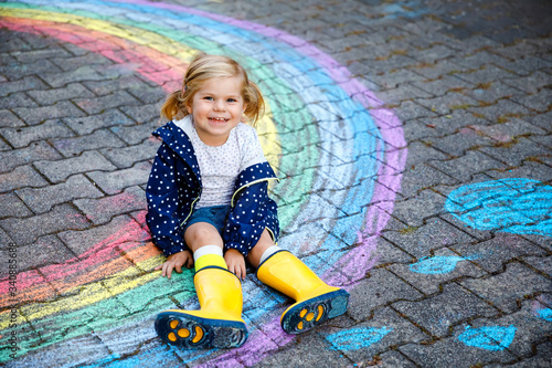 Happy little toddler girl in rubber boots with rainbow painted with colorful chalks on ground during pandemic coronavirus quarantine. Children painting rainbows along with the words Let's all be well photo