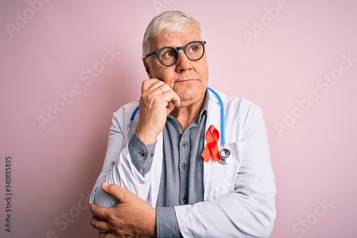 Senior handsome hoary doctor man wearing stethoscope and red HIV ribbon on coat with hand on chin thinking about question, pensive expression. Smiling with thoughtful face. Doubt concept. © Krakenimages.com