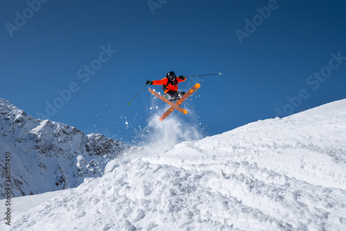 Male athlete skier in an orange trigger makes a jump trick with flying snow powder against the backdrop of snow-capped Caucasus mountains and a blue sky. Winter Extreme Sports Concept