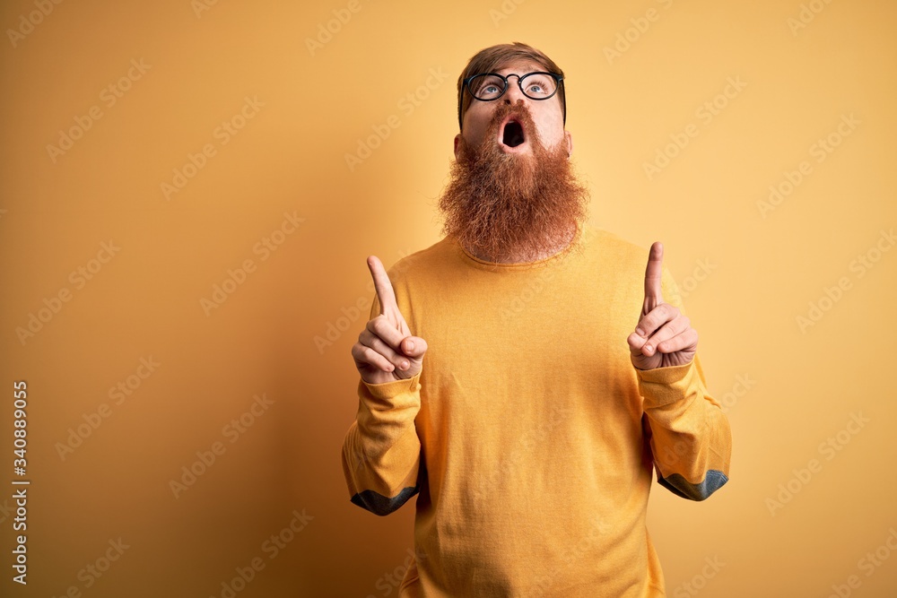Handsome Irish redhead man with beard wearing glasses over yellow isolated background amazed and surprised looking up and pointing with fingers and raised arms. Handsome Irish redhead man with beard wearing glasses over yellow isolated background amazed and surprised looking up and pointing with fingers and raised arms.