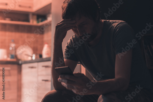 Depressed man typing text message on mobile phone in dark room