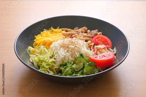 Traditional Korean dish Kuksi on a wooden background (one of the types of Kuksi). Ingredients -pasta, vegetable salads of cucumbers, cabbage, radish, meat, omelet. The broth is served separately.