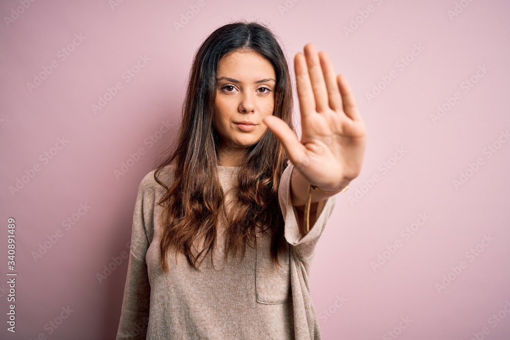 Young beautiful brunette woman wearing casual sweater standing over pink background doing stop sing with palm of the hand. Warning expression with negative and serious gesture on the face. Young beautiful brunette woman wearing casual sweater standing over pink background doing stop sing with palm of the hand. Warning expression with negative and serious gesture on the face.