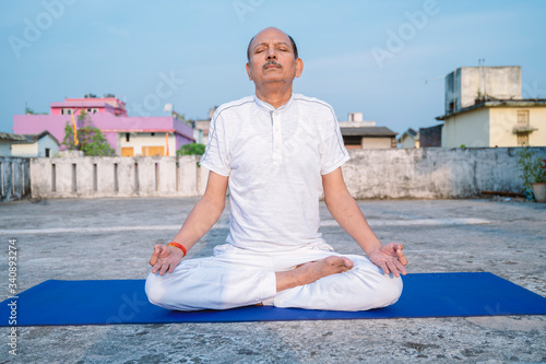 Senior Citizen or an Old Indian Man Performing Yoga Early Morning, in his Terrace in white Tshirt and Pants. Stay Home Stay Safe and Fit