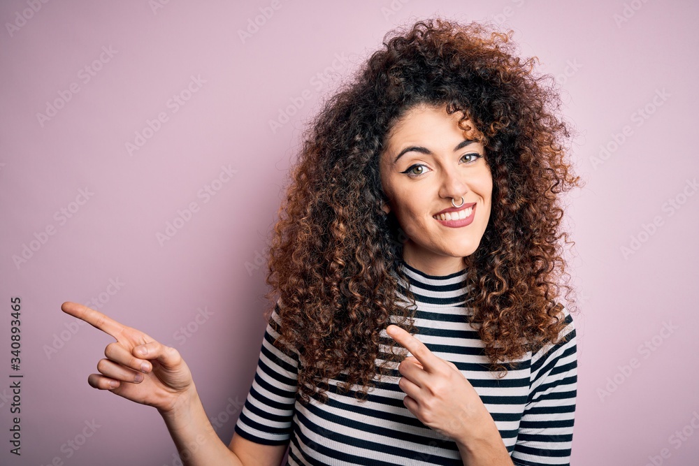 Young beautiful woman with curly hair and piercing wearing casual striped t-shirt smiling and looking at the camera pointing with two hands and fingers to the side. Young beautiful woman with curly hair and piercing wearing casual striped t-shirt smiling and looking at the camera pointing with two hands and fingers to the side.