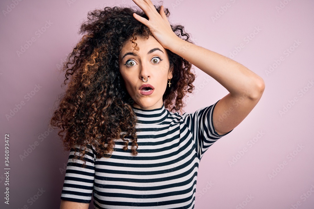 Young beautiful woman with curly hair and piercing wearing casual striped t-shirt surprised with hand on head for mistake, remember error. Forgot, bad memory concept. Young beautiful woman with curly hair and piercing wearing casual striped t-shirt surprised with hand on head for mistake, remember error. Forgot, bad memory concept.