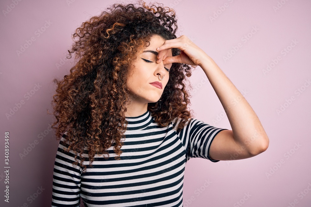 Young beautiful woman with curly hair and piercing wearing casual striped t-shirt tired rubbing nose and eyes feeling fatigue and headache. Stress and frustration concept. Young beautiful woman with curly hair and piercing wearing casual striped t-shirt tired rubbing nose and eyes feeling fatigue and headache. Stress and frustration concept.
