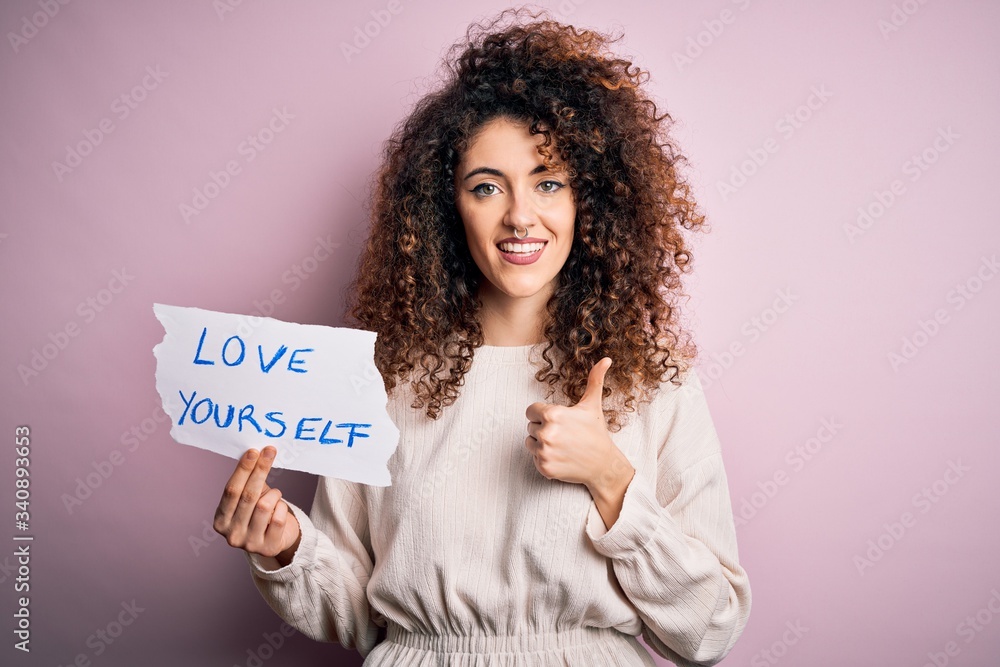 Young beautiful woman with curly hair and piercing holding paper with love yourself message happy with big smile doing ok sign, thumb up with fingers, excellent sign Young beautiful woman with curly hair and piercing holding paper with love yourself message happy with big smile doing ok sign, thumb up with fingers, excellent sign