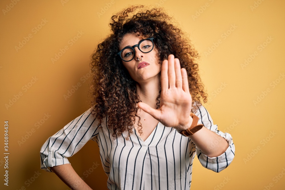 Young beautiful woman with curly hair and piercing wearing striped shirt and glasses doing stop sing with palm of the hand. Warning expression with negative and serious gesture on the face. Young beautiful woman with curly hair and piercing wearing striped shirt and glasses doing stop sing with palm of the hand. Warning expression with negative and serious gesture on the face.