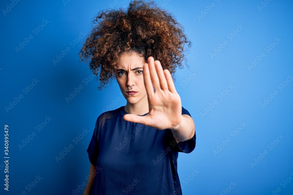 Young beautiful woman with curly hair and piercing wearing casual blue t-shirt doing stop sing with palm of the hand. Warning expression with negative and serious gesture on the face. Young beautiful woman with curly hair and piercing wearing casual blue t-shirt doing stop sing with palm of the hand. Warning expression with negative and serious gesture on the face.